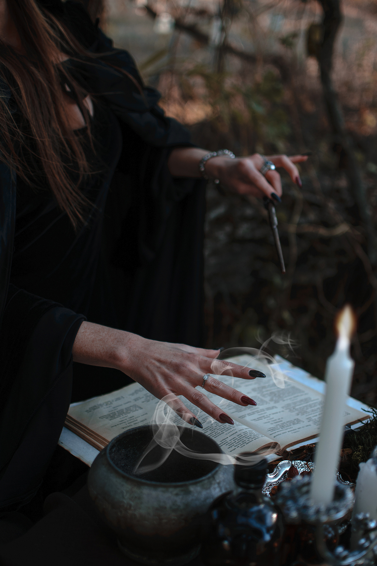 A Woman in Black Robe Holding a Stick Near the Table with Book and Candle